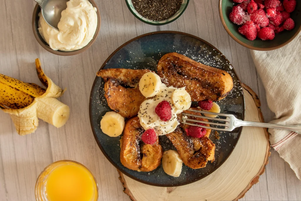 Golden-brown Hawaiian Roll French Toast topped with powdered sugar, fresh berries, and banana on a rustic wooden table.